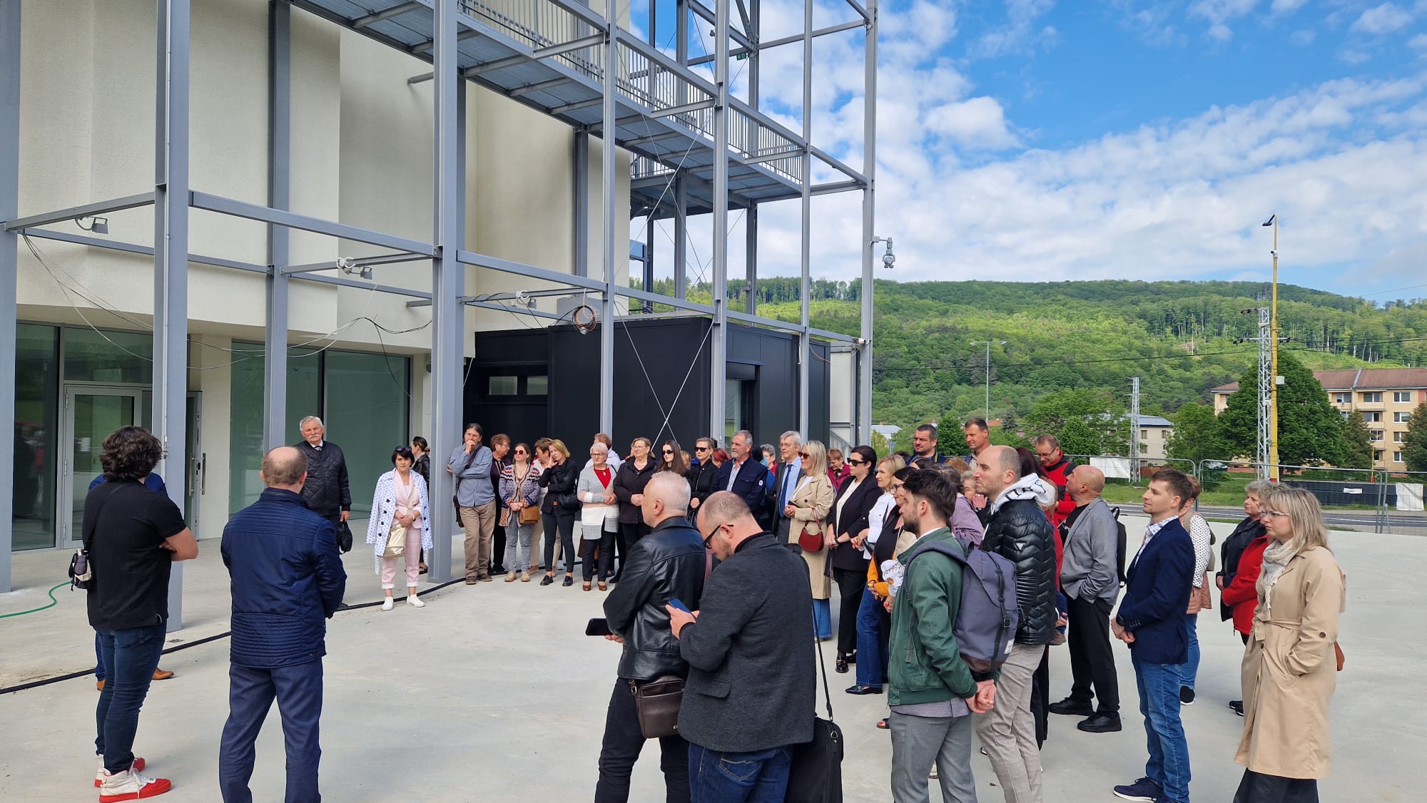 Grupa ludzi zebrała się przed nowoczesnym budynkiem z metalową konstrukcją, zalesione wzgórza i błękitne niebo w tle.
